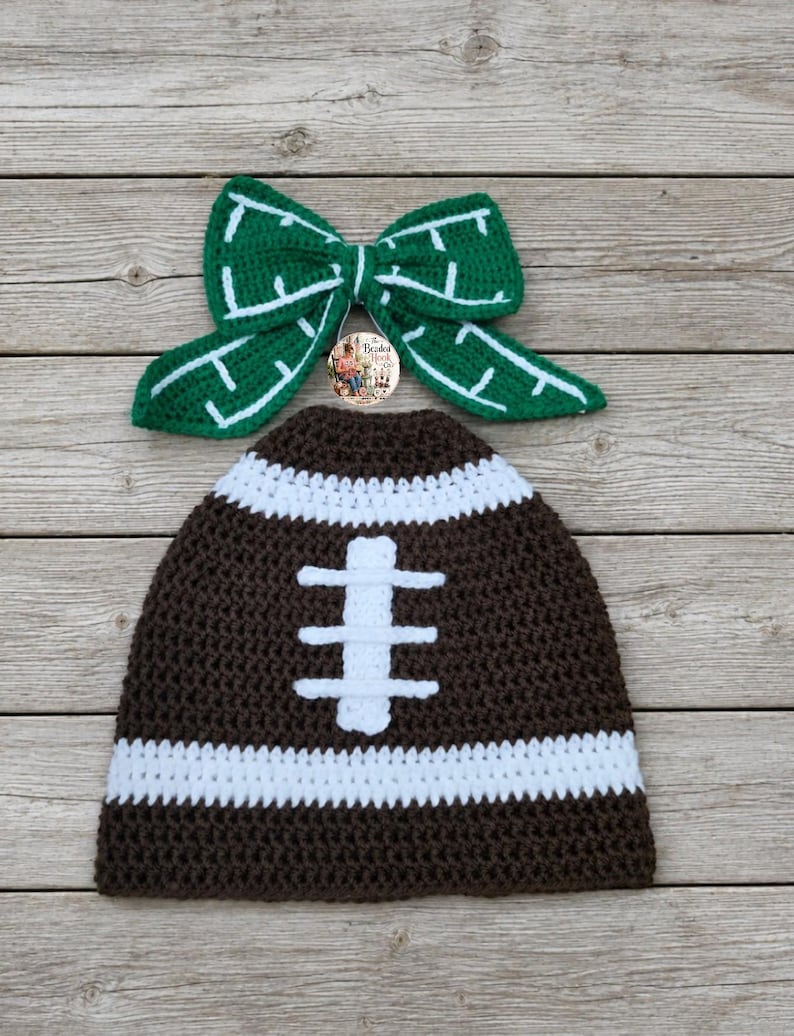 May include: A dark brown crochet beanie with a white horizontal stripe and a white vertical stitch pattern resembling a football, paired with a green crochet bow featuring white stitched lines and a small circular logo tag in the center.