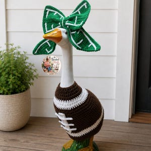 May include: A white goose statue wearing a brown and white crocheted football sweater and a green and white crocheted bow headband. The sweater features white laces and stripes. A circular logo with the text The Beaded Hook Co. is visible.