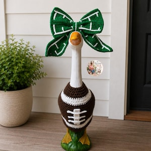 May include: A white decorative goose statue wearing a brown and white crocheted football sweater and a large green and white crocheted bow on its head. The sweater features white laces and stripes, while the bow has a white grid pattern.