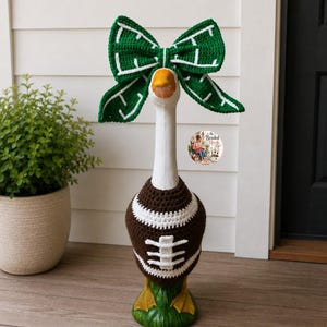 May include: A white decorative goose statue wearing a brown and white crocheted football sweater and a large green and white crocheted bow on its head. The sweater features white laces and stripes, while the bow has a white grid pattern.
