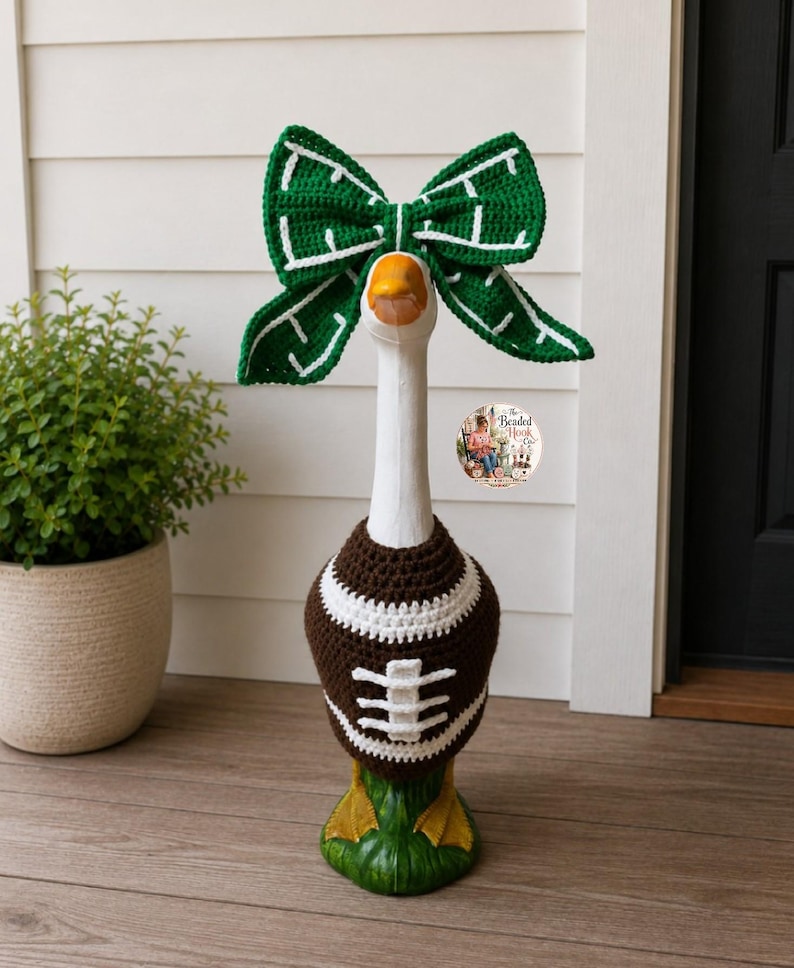 May include: A white decorative goose statue wearing a brown and white crocheted football sweater and a large green and white crocheted bow on its head. The sweater features white laces and stripes, while the bow has a white grid pattern.