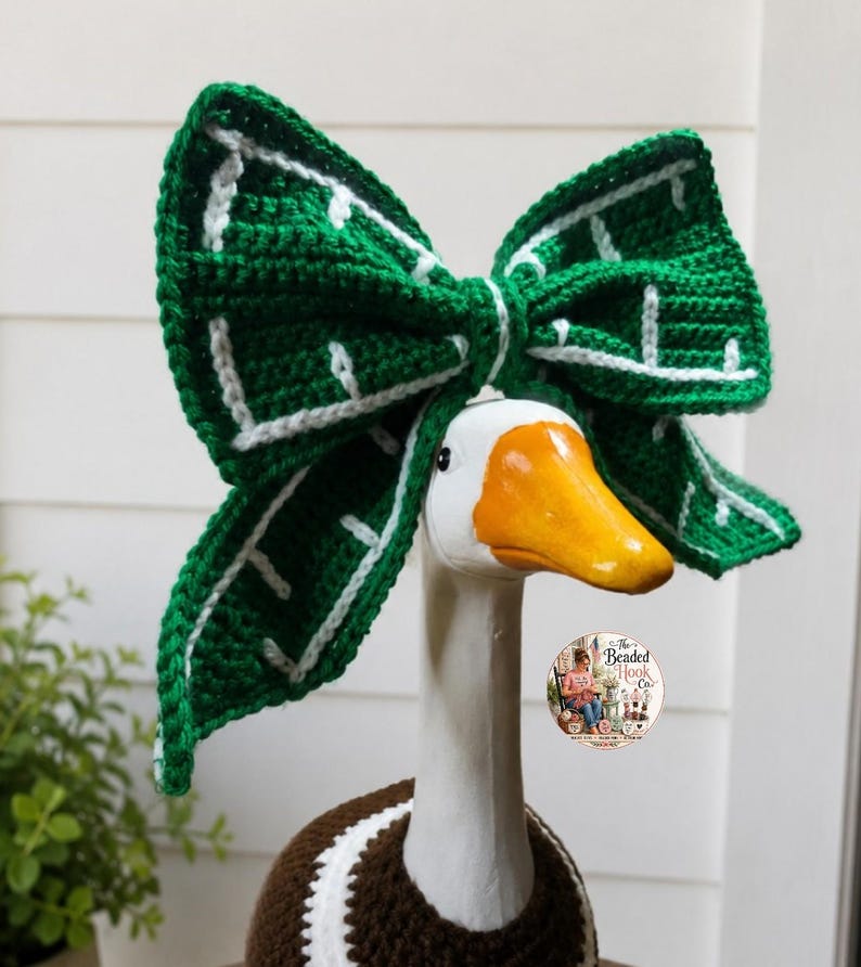 May include: A white goose figurine wearing a large, crocheted green bow with white grid-like accents on its head. The goose also wears a brown and white crocheted garment around its neck. A circular logo with text is visible on the neck area.