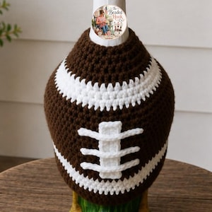 May include: A brown and white crocheted cover shaped like an American football. The cover features a dark brown textured body with white horizontal stripes at the top and bottom, and a central white vertical lace detail resembling football stitching.