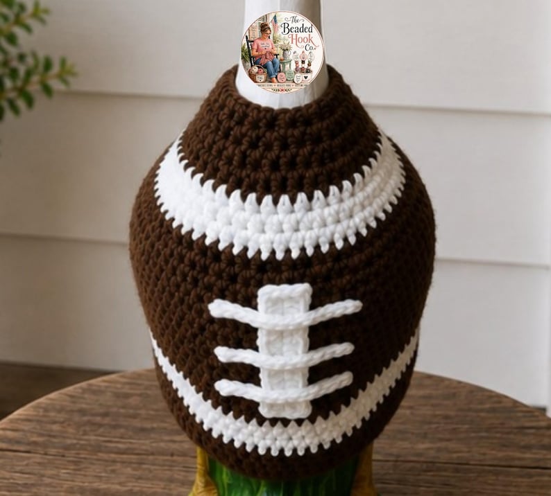 May include: A brown and white crocheted cover shaped like an American football. The cover features a dark brown textured body with white horizontal stripes at the top and bottom, and a central white vertical lace detail resembling football stitching.