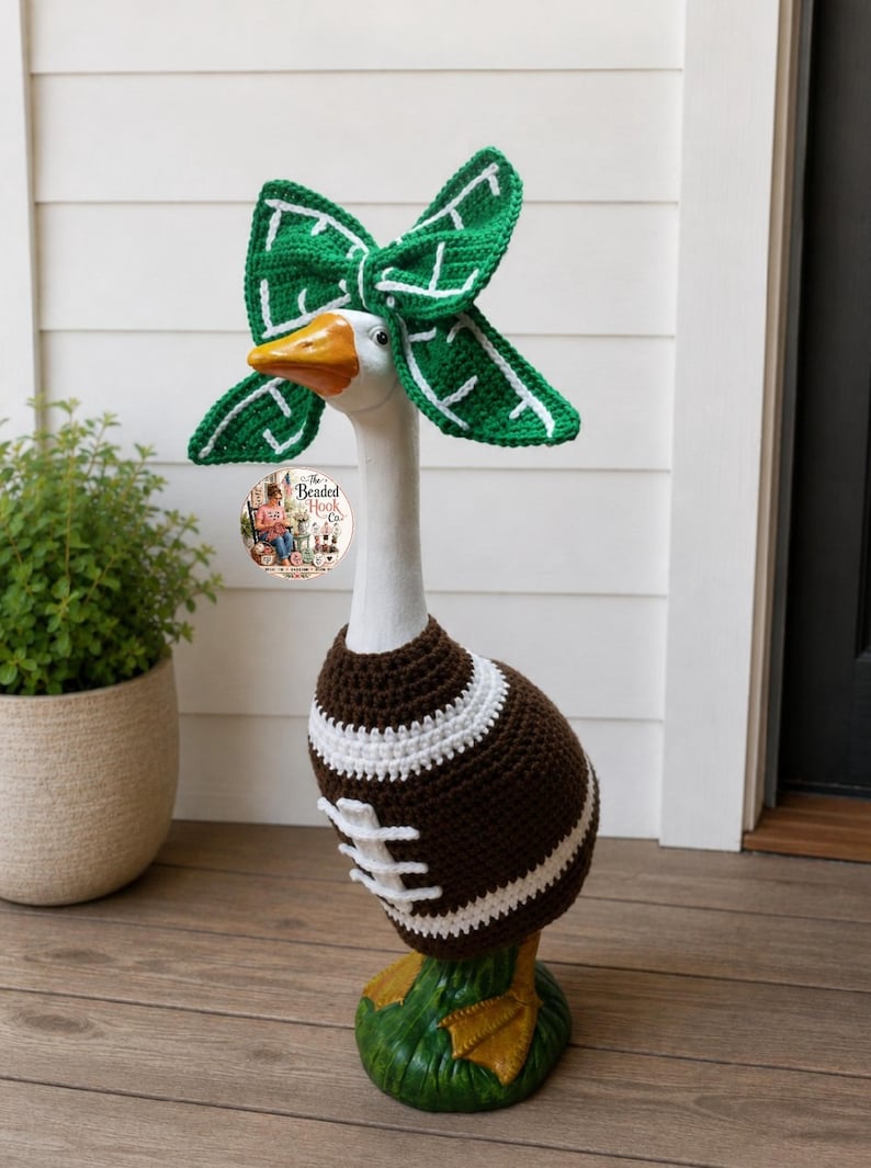 May include: A white goose statue wearing a brown and white crocheted football sweater and a green and white crocheted bow headband. The sweater features white laces and stripes. A circular logo with the text The Beaded Hook Co. is visible.