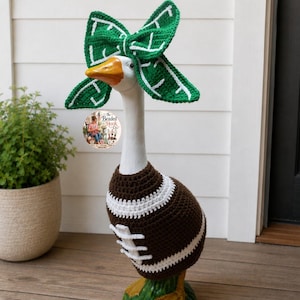 May include: A white goose statue wearing a brown and white crocheted football sweater and a green and white crocheted bow headband. The sweater features white laces and stripes. A circular logo with the text The Beaded Hook Co. is visible.