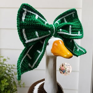 May include: A white goose figurine wearing a large, crocheted green bow with white grid-like accents on its head. The goose also wears a brown and white crocheted garment around its neck. A circular logo with text is visible on the neck area.