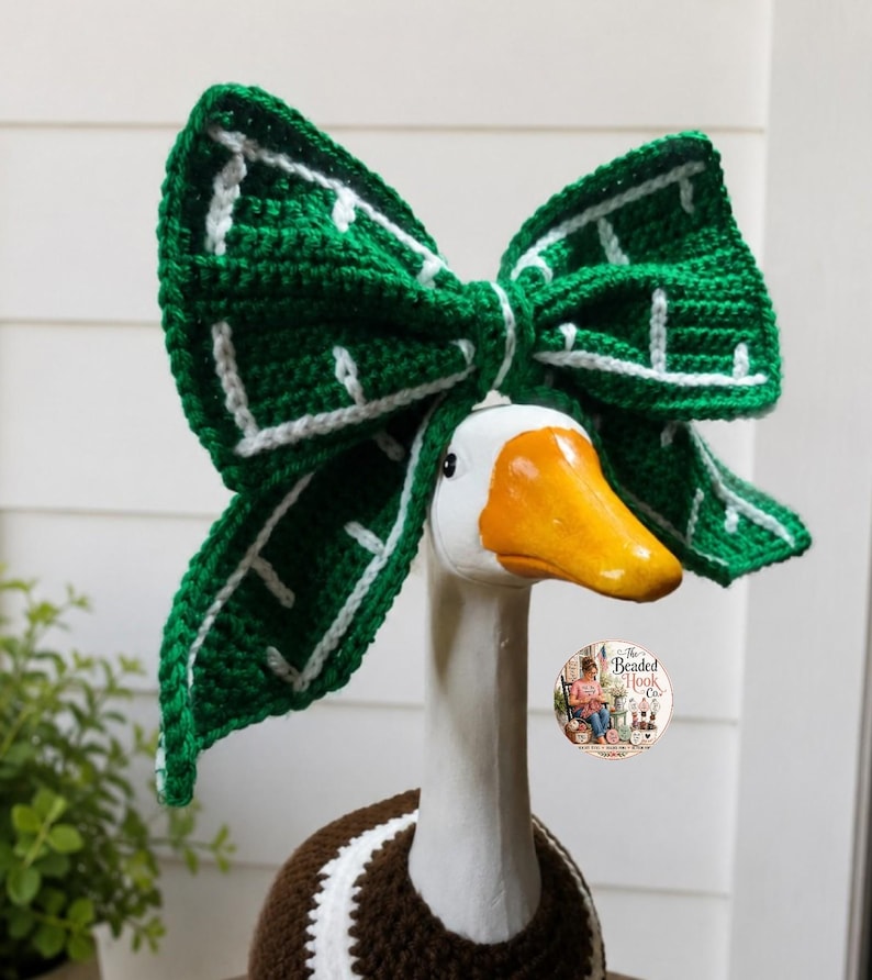 May include: A white goose figurine wearing a large, crocheted green bow with white grid-like accents on its head. The goose also wears a brown and white crocheted garment around its neck. A circular logo with text is visible on the neck area.