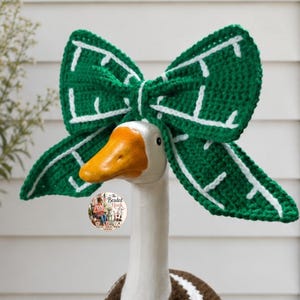 May include: A large, dark green crochet bow accessory with white stitched leaf-like vein patterns, positioned atop a white goose figurine. The bow features a central knot and wide, pointed wings. A small circular logo is visible on the goose's neck.