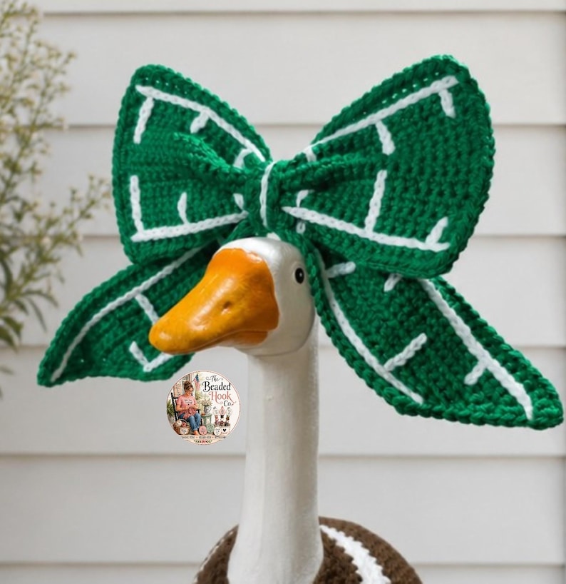 May include: A large, dark green crochet bow accessory with white stitched leaf-like vein patterns, positioned atop a white goose figurine. The bow features a central knot and wide, pointed wings. A small circular logo is visible on the goose's neck.