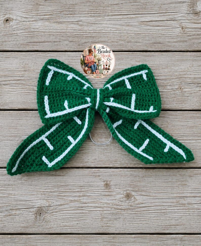 May include: A dark green crochet bow featuring a white stitched pattern that resembles a maze or grid design. The bow is constructed with a central knot and two long, pointed tails, and it includes a circular metal ring attached to the back.