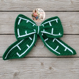 May include: A dark green crochet bow featuring a white stitched pattern that resembles a maze or grid design. The bow is constructed with a central knot and two long, pointed tails, and it includes a circular metal ring attached to the back.