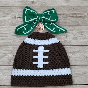 May include: A dark brown crochet beanie with a white horizontal stripe and a white vertical stitch pattern resembling a football, paired with a green crochet bow featuring white stitched lines and a small circular logo tag in the center.