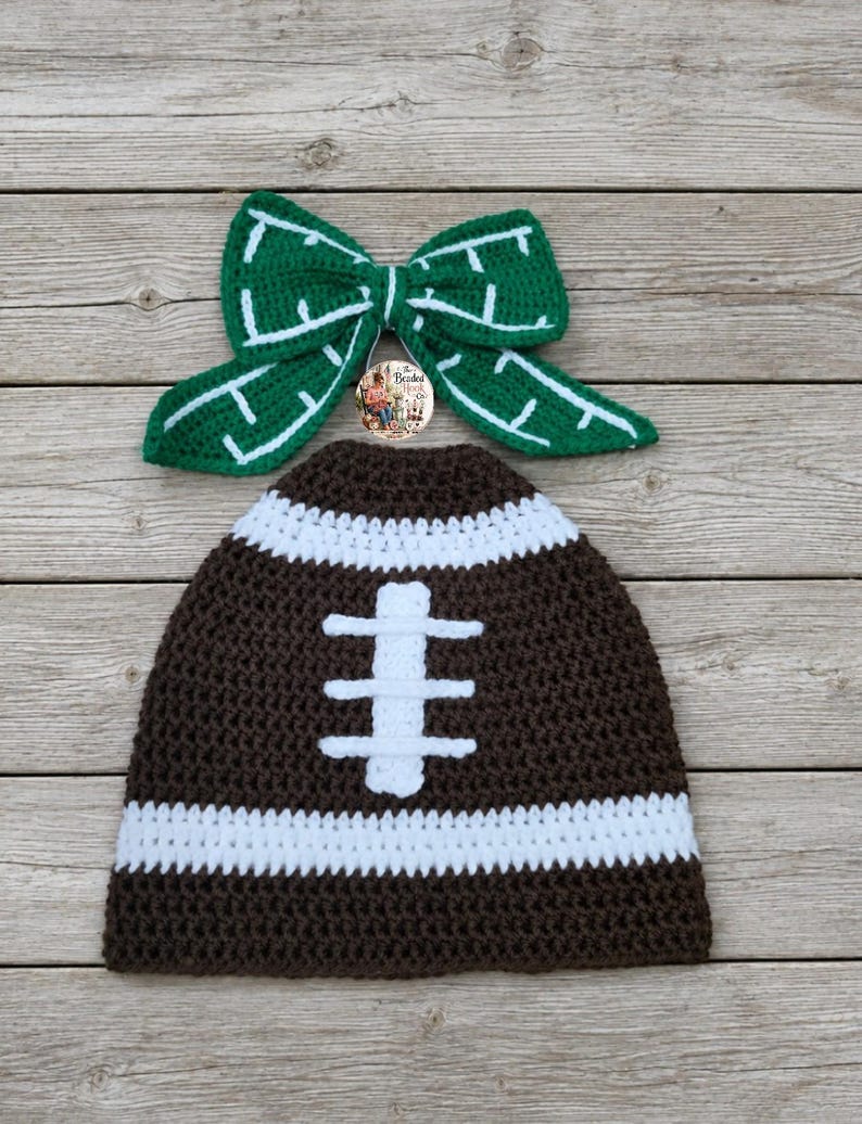 May include: A dark brown crochet beanie with a white horizontal stripe and a white vertical stitch pattern resembling a football, paired with a green crochet bow featuring white stitched lines and a small circular logo tag in the center.