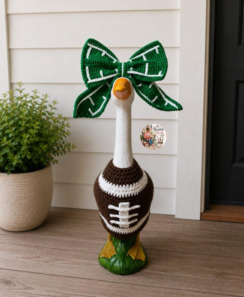 May include: A white decorative goose statue wearing a brown and white crocheted football sweater and a large green and white crocheted bow on its head. The sweater features white laces and stripes, while the bow has a white grid pattern.