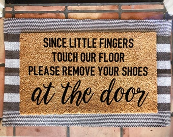 May include: A brown coir doormat with white and gray stripes around the edges. The doormat has the text "Since little fingers touch our floor please remove your shoes at the door" in black lettering.