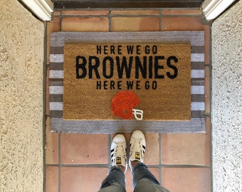 May include: A brown doormat with white stripes and the text "Here we go Browns Here we go" in black. An orange and black football helmet is centred on the mat. A pair of white trainers are on the floor in front of the mat.
