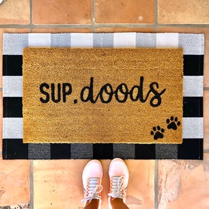 May include: A brown coir doormat with the text "SUP. doods" in black cursive lettering and two black paw prints. The doormat is on top of a black and white checkered rug.