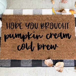 May include: A rectangular brown coir doormat with the text "HOPE YOU BROUGHT pumpkin cream cold brew" in black lettering. The mat is on a black and white checkered rug, with fall leaves and decorative pumpkins.