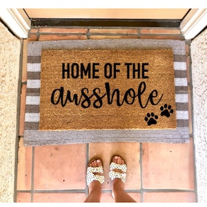 May include: A brown coir doormat with the text "Home of the Asshole" in black cursive lettering. Three black paw prints are on the right side of the mat. The mat is on a tiled floor with a white and gray striped border.