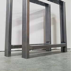 May include: Two metal table legs with a rectangular frame and a single crossbar. The legs are made of steel and have a dark gray finish.