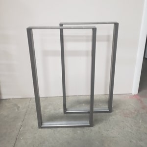 May include: Two metal table legs in a rectangular shape, made of steel, with a gray finish. The legs are standing on a concrete floor.