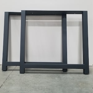 May include: Two black metal table legs with a rectangular top and a slightly angled design. The legs are standing on a concrete floor.