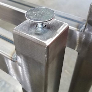 May include: Close-up of a silver metal weld-on cap with an adjustable leveling foot. The cap is attached to a square metal leg.