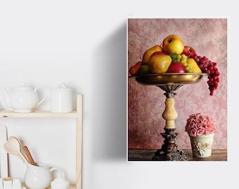 May include: A gold metal fruit bowl with a pedestal base, filled with red apples, yellow pears, and green grapes. The bowl is on a pink patterned background with a pink flower arrangement in a white ceramic vase.