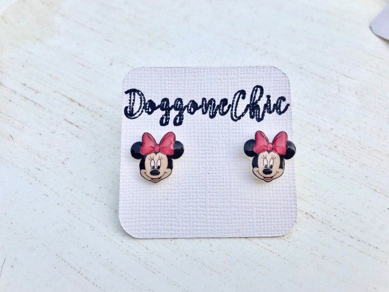 Puede incluir: Un par de pendientes de bot&oacute;n de Minnie Mouse en una tarjeta blanca con el texto "Doggone Chic". Los pendientes son negros con un lazo rojo y una cara blanca.