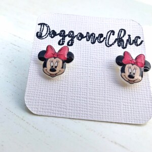 Puede incluir: Un par de pendientes de bot&oacute;n de Minnie Mouse con fondo blanco y lazos rojos. Los pendientes est&aacute;n en una tarjeta blanca con el texto "Doggone Chic" escrito en negro.