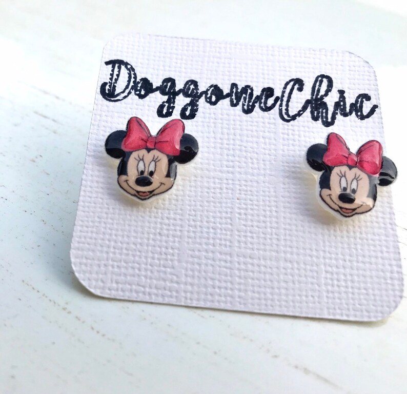 Puede incluir: Un par de pendientes de bot&oacute;n de Minnie Mouse con fondo blanco y lazos rojos. Los pendientes est&aacute;n en una tarjeta blanca con el texto "Doggone Chic" escrito en negro.