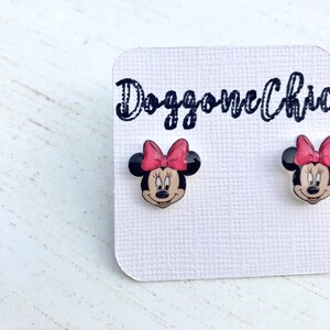 Puede incluir: Un par de pendientes de bot&oacute;n de Minnie Mouse con un lazo rojo y orejas negras. Los pendientes est&aacute;n en una tarjeta blanca con el texto "Doggone Charmed" en negro.