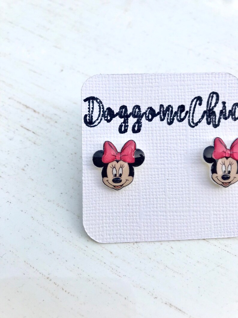 Puede incluir: Un par de pendientes de bot&oacute;n de Minnie Mouse con un lazo rojo y orejas negras. Los pendientes est&aacute;n en una tarjeta blanca con el texto "Doggone Charmed" en negro.