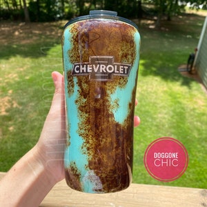 May include: A turquoise and brown stainless steel tumbler with a rusty effect and a silver Chevrolet logo. The tumbler is held in a hand and the background is a blurry green lawn.