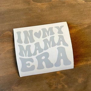 May include: A white sticker with the text "In My Mama Era" in a retro font. The text is in grey and the heart is a light pink.