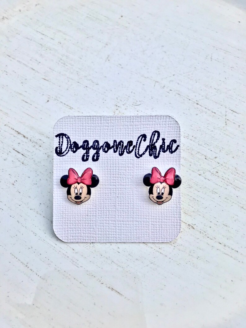 Puede incluir: Un par de pendientes de bot&oacute;n de Minnie Mouse con un lazo rosa en una tarjeta blanca con el texto "Doggone Chic".