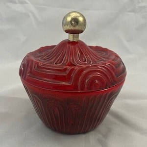 May include: A red ceramic trinket box with a gold-colored knob on the lid. The box has a geometric design.