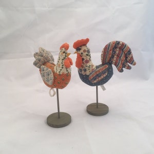 May include: Two colorful fabric roosters on stands. The rooster on the left is orange, green, and blue. The rooster on the right is blue, orange, and red.