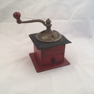May include: Antique red wooden coffee grinder with a metal crank and a drawer. The grinder has a black metal top with a decorative design.
