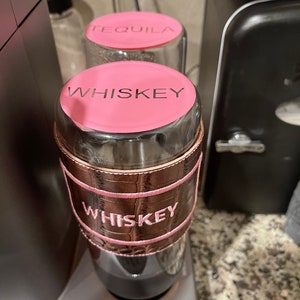 May include: Two glass bottles with pink lids and brown leather sleeves. The lids are labeled "Tequila" and "Whiskey".