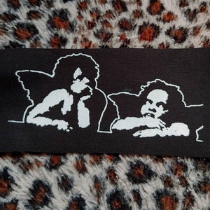 以下が含まれることがあります： 黒と白の刺繍パッチで、翼のある天使が2人描かれています。一人は横を向いており、もう一人は正面を見ています。