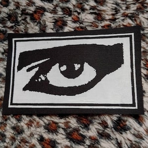 May include: Black and white embroidered patch with a stylized eye design. The eye is outlined in white and has a black pupil. The patch is surrounded by a white border.