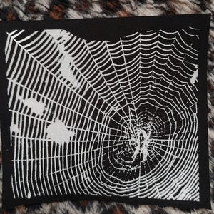 May include: Black fabric patch featuring a white spiderweb design. The web is detailed and intricate, with a central point where the web radiates outwards.