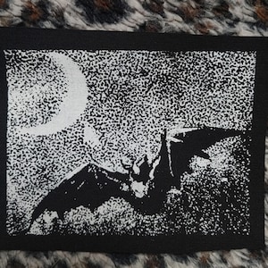 May include: A black and white patch depicting a bat flying in front of a crescent moon and a starry sky. The patch has a black border. The design is a stylized illustration, possibly for clothing or accessories.