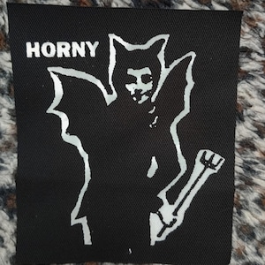 May include: Black iron-on patch with a white silhouette of a winged creature holding a weapon. The text "HORNY" is printed in white at the top.