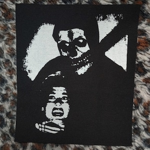 May include: Black and white embroidered patch featuring a skull-faced figure holding a child. The patch is on a black fabric background.