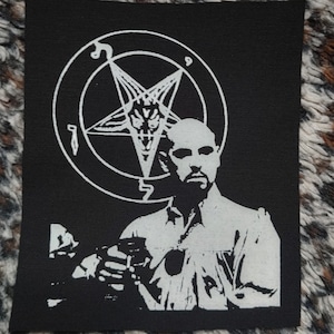 Puede incluir: Un parche blanco y negro con un pentagrama y un hombre sosteniendo un vaso. El pentagrama tiene una cabeza de cabra en el centro. El hombre lleva una camisa blanca y tiene una expresión seria.