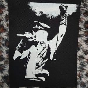 Könnte beinhalten: Schwarz-weißes Bügeleisen-Patch mit der Silhouette einer Person, die ein Mikrofon und eine Gitarre hält, mit erhobenem Arm.
