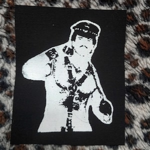 May include: A black and white patch with a person wearing a hat and leather clothing. The person is standing with their arm raised and their hand in a fist.